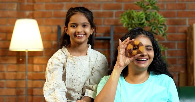Indian Asian young mother and cute little daughter sitting on a sofa in a lavish modern home, playfully holding a round donut, peeping through the hole, smiling and making an okay hand gesture