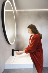 Young woman in a red robe washing her hands under running water at a modern bathroom sink with a...