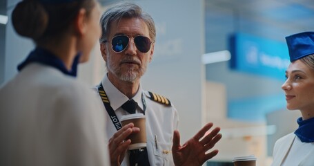 Airline Captain in Glasses Talks to Two Beautiful Stewardesses in Uniforms, Drinks Coffee, Waits for Commercial Flight. Flight Crew in Crowded International Airport Terminal. Close Up. Slow Motion