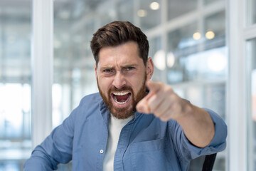An angry man in a blue shirt points directly at the viewer, yelling with an open mouth and fierce...