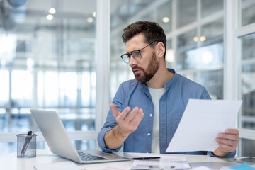 A businessman wearing glasses is working with papers and laptop, looking confused or frustrated while in an office environment.