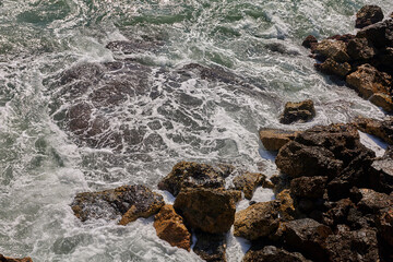 Foamy waves crash against dark wet rocks with patches of yellow and brown tones under sunlight reflections; tourism;