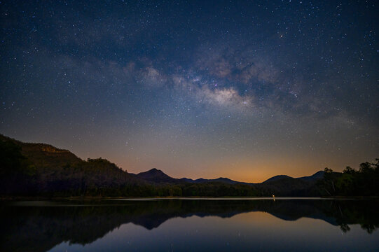 milky way stretches across the star-filled night sky above dark mountain silhouettes and a calm reflective lake, with a soft orange glow near the horizon