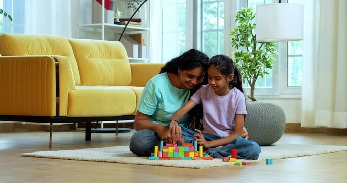 Indian Asian young mother and small daughter happily building a house using colorful wooden block toys while sitting together on carpeted floor of a modern lavish home, spending quality time indoors