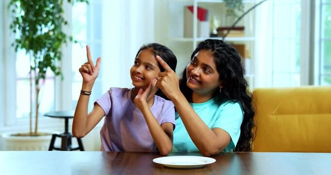 Indian Asian young woman and cute little daughter with empty white plate sitting at dining table in modern home, showing tasty gesture with hand while pointing at plate, holding fork, spoon playfully