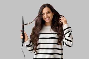 Beautiful young woman with curling iron on grey background