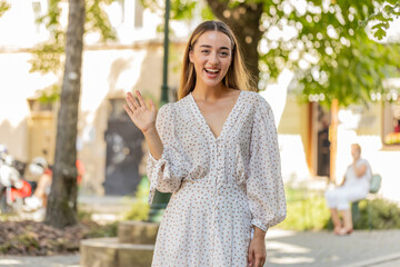 Hello. Caucasian young woman in long dress smiling friendly at camera, waving hands gesturing hi greeting or goodbye welcoming with hospitable expression outdoors. Girl standing in urban city street. © Andrii Iemelianenko