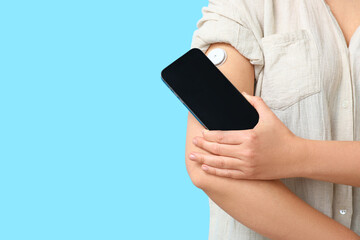 Woman with glucose sensor for measuring blood sugar level and phone on blue background, closeup....