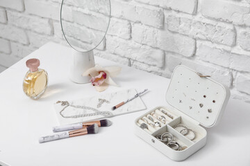 Box with stylish different jewelry, mirror and perfume bottle on dresser near white brick wall