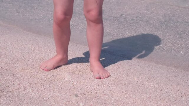 Child's bare feet stand on sandy beach at water's edge, casting a distinct shadow in the sunlight. The scene captures the simplicity of a seaside moment