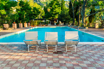 three white desk chairs or chaise lounge with blue swimming pool and hotel green garden on backgeound of vacation summer landscape