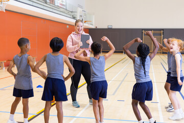 In school gym, female coach using tablet to instruct diverse children during fitness class