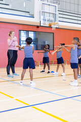 Fototapeta premium In school gym, diverse children exercising with female teacher, stretching arms in group activity