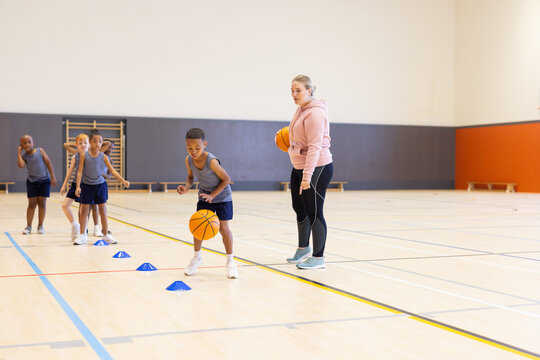 In school gym, diverse children practicing basketball drills with female coach supervising
