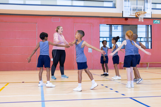 In school gym, diverse children playing game with female teacher, holding hands in circle