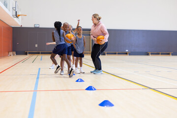 Playing basketball, diverse children in school gym practicing with female coach and cones