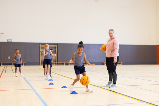 In school gym, diverse children practicing basketball drills with female coach