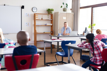 In school, female teacher sitting at desk, engaging with diverse students in classroom