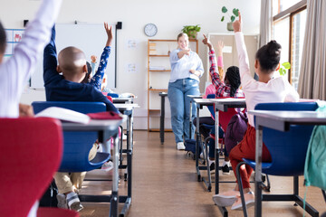 In school, diverse students raising hands while female teacher pointing in classroom