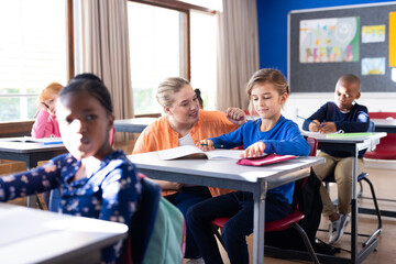 Helping boy with homework, female teacher in school classroom, other students studying