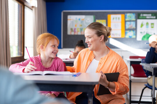 In school, female teacher helping girl with homework at desk, smiling together