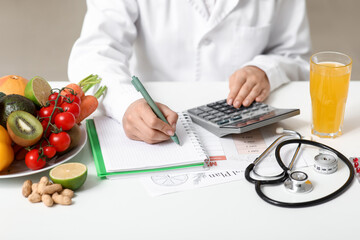 Young female nutritionist with healthy food, glass of juice and calculator writing diet plan in...