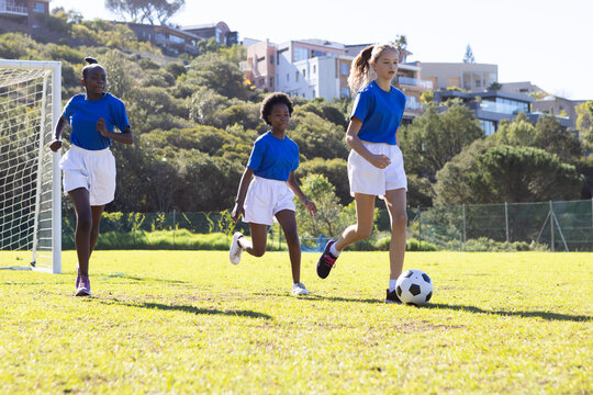 Fototapeta Playing soccer on school field, diverse girls in sports uniforms running with ball