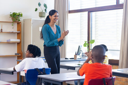Indian female teacher clapping hands while students sitting at desks in school classroom