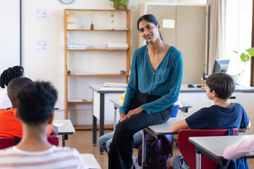 In school, Indian female teacher sitting on desk and engaging with students in classroom
