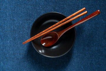 A pair of wooden chopsticks and spoon lying across an empty black soup bowl on blue fabric...
