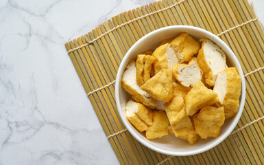 Close-up of deep-fried tofu in a white bowl on a bamboo mat on a white marble table. Flat-lay food image with space.
