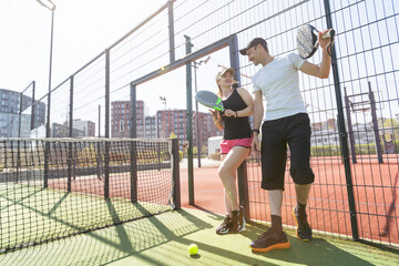 Portrait of positive young woman and adult man standing on padel tennis court, holding racket and ball, smiling 