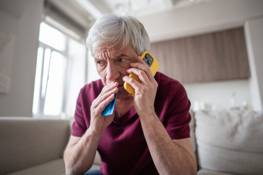 Nervous pensioner man hesitates with payment card in hand, hanging on phone, voice demands immediate account access. Social engineering, fraudulent purchase attempt, vulnerable senior male target 