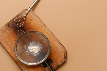 Metal sieve with flour, wooden board and whisk on color background