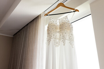 Elegant wedding dress hanging by window, showcasing intricate lace details, at home, copy space