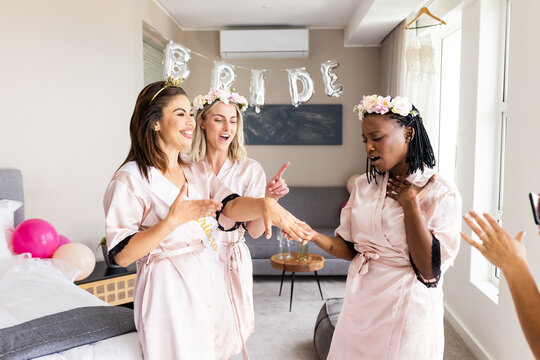 Celebrating at home, diverse friends in robes admiring engagement ring with joy