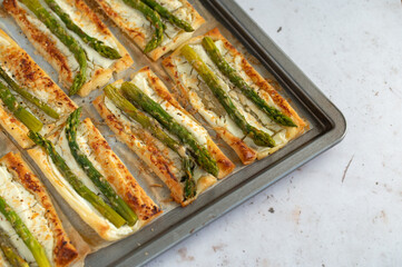 Small puff pastry tarts with green asparagus, fresh cream and parmesan. Asparagus pie with cheese on a metal tray on stone background. Spring healthy food.