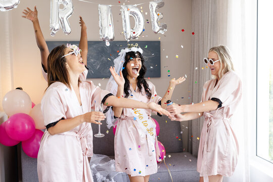Diverse female friends in robes, confetti, laughing at bachelorette party celebration, at home