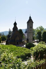 Vang Stave Church and its stone tower stand prominently amidst green vegetation under a clear sky