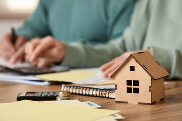 House figure on table against senior couple calculating utility bills in kitchen, closeup