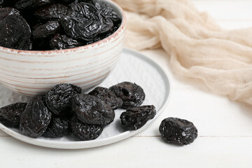 Bowl and plate with dried prunes on white wooden background