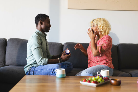 African American man proposing to surprised woman on sofa, holding engagement ring, copy space