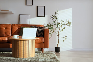 Interior of living room with houseplant, stylish leather sofa and laptop on table near white wall