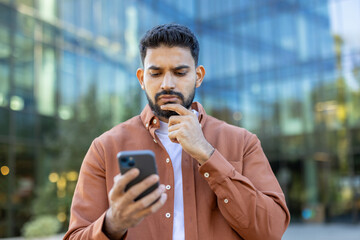 A man considers his phone in front of a modern building, contemplating the content on the screen. Thinking about communication.