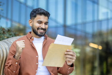 A young man celebrates with excitement, revealing a positive letter result with a clenched fist and a big smile, outdoors.
