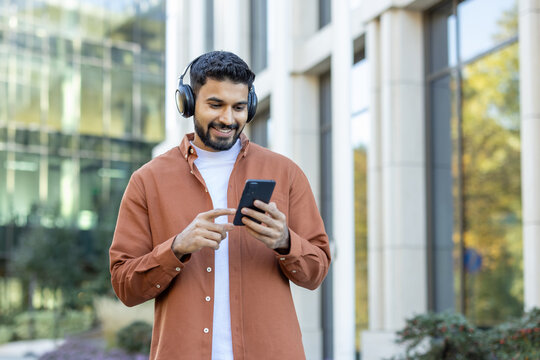 A smiling man in headphones checks his phone outdoors, near a modern building and greenery. He appears to be reading a message.