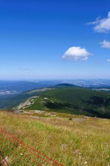 A vast mountain landscape with grassy slopes, a winding trail with hikers, and distant hazy views under a blue sky with scattered clouds