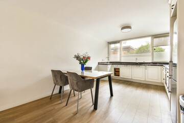 A bright and modern kitchen interior featuring a dining table, chairs, and white cabinetry. Natural light fills the space through large windows.