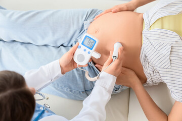 Young pregnant woman on couch and doctor with fetal doppler listening to baby's heartbeat in clinic, top view