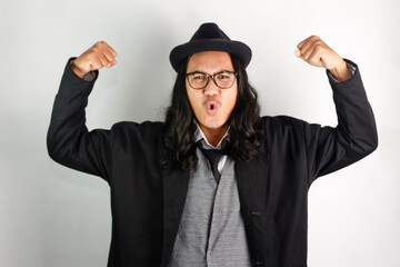 Asian man with long hair dressed as a mafia wearing glasses and a hat, making an exited expression, both hands making muscle-showing gestures
